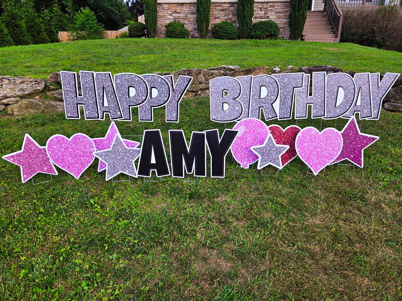 Example of a birthday greeting that reads 'Happy Birthday Amy' with glittery text and decorative balloon, star, and heart signs in shades of pink